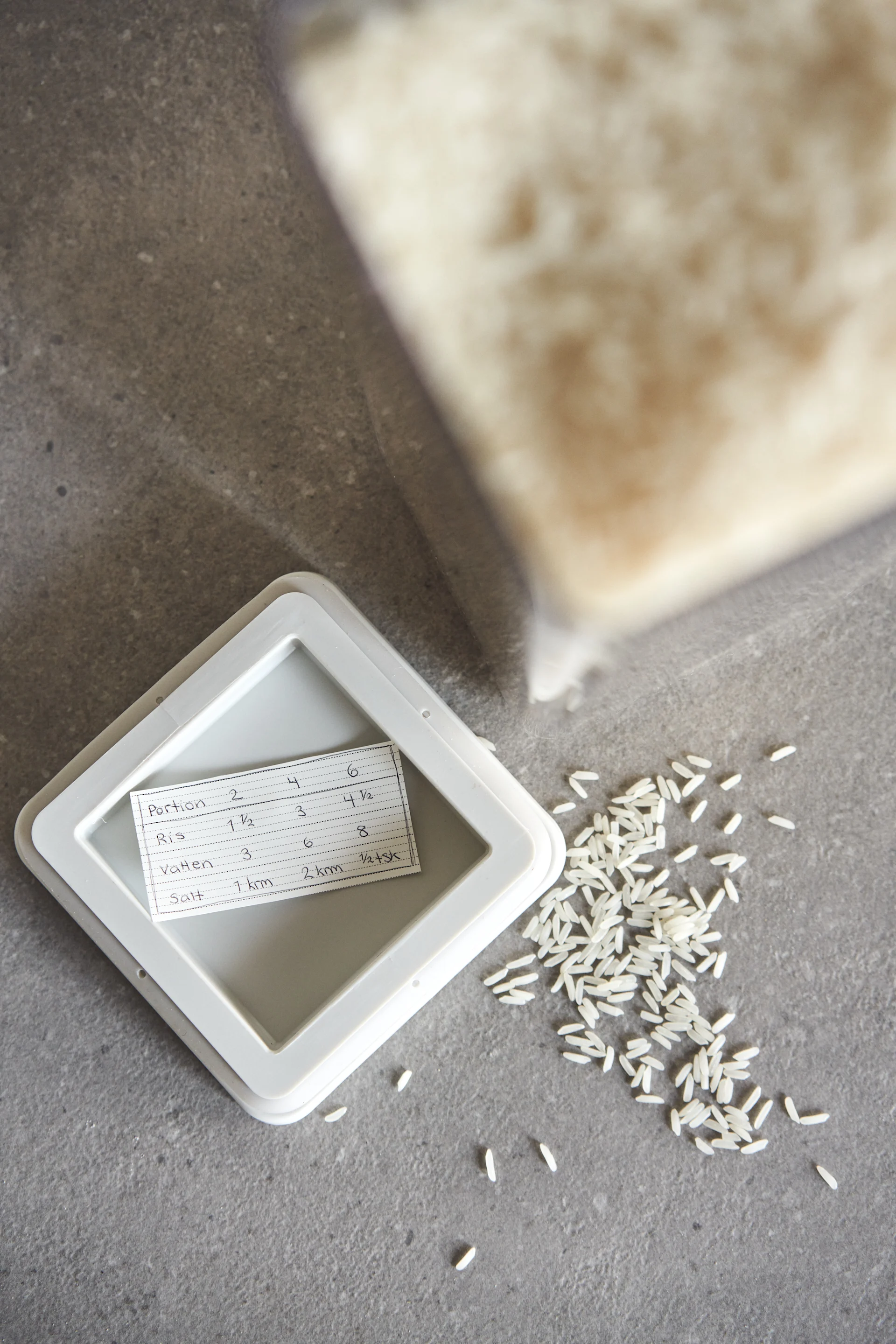 Recipe card for rice on a white lid, next to scattered grains and a blurred bag of rice.