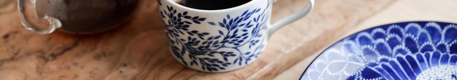White mug with blue leaf pattern and coffee on a wooden table next to a blue patterned plate.