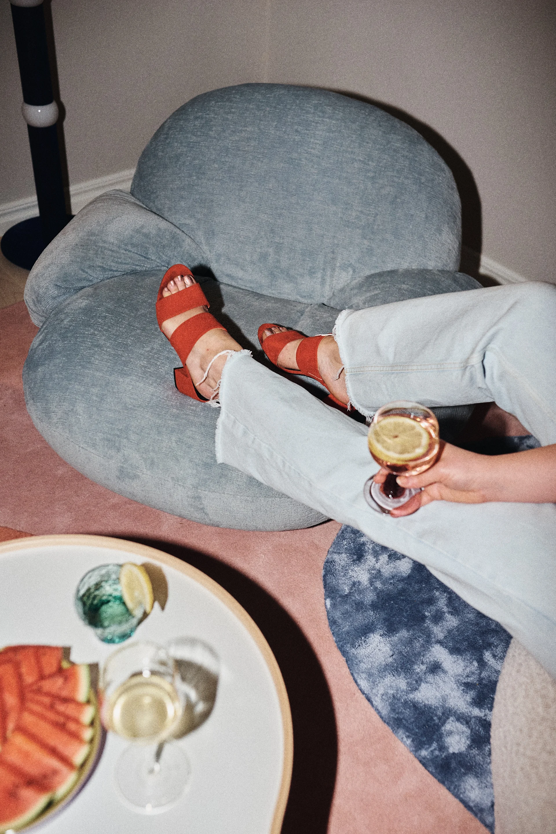 Person lounging on a blue bean bag chair, wearing light jeans and red sandals, holding a drink with a lemon. Sliced watermelon and another drink are on a table nearby.