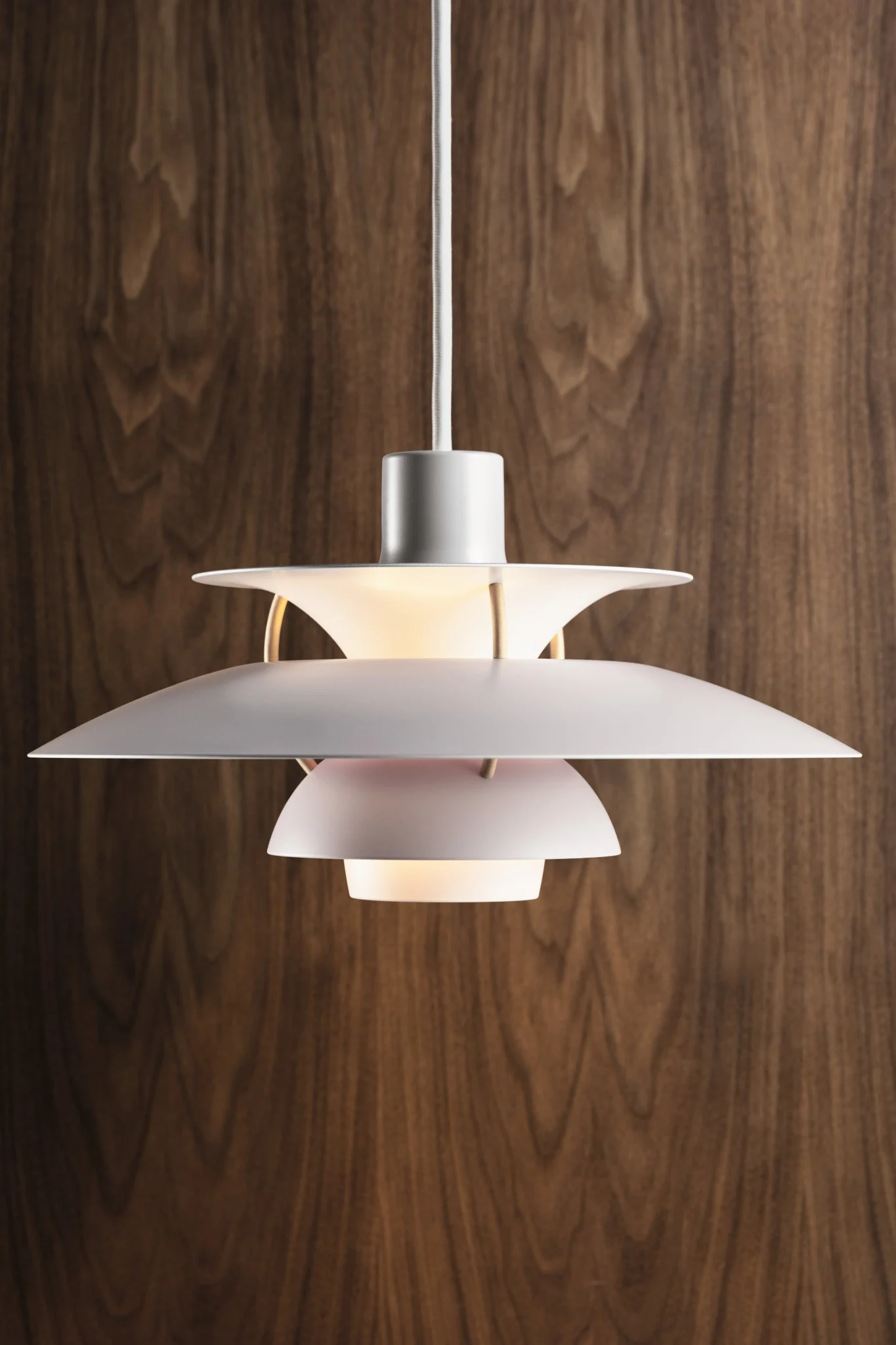 A white multi-tiered pendant lamp with a warm glow, hanging against a dark wood panel wall.
