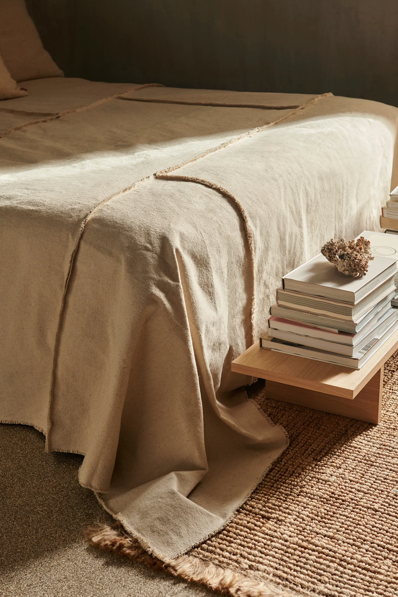 Bed with natural textured blanket, stack of books, coral on wooden table, and woven rug.
