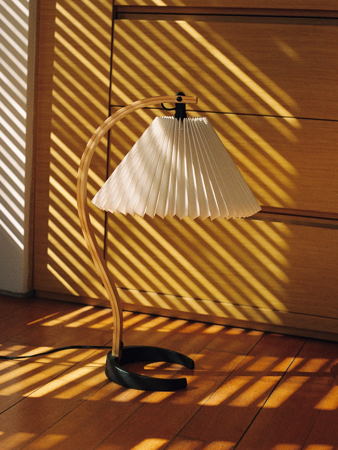Desk lamp with a curved wooden stand and pleated shade, illuminated by diagonal sunlight stripes on a wooden floor.