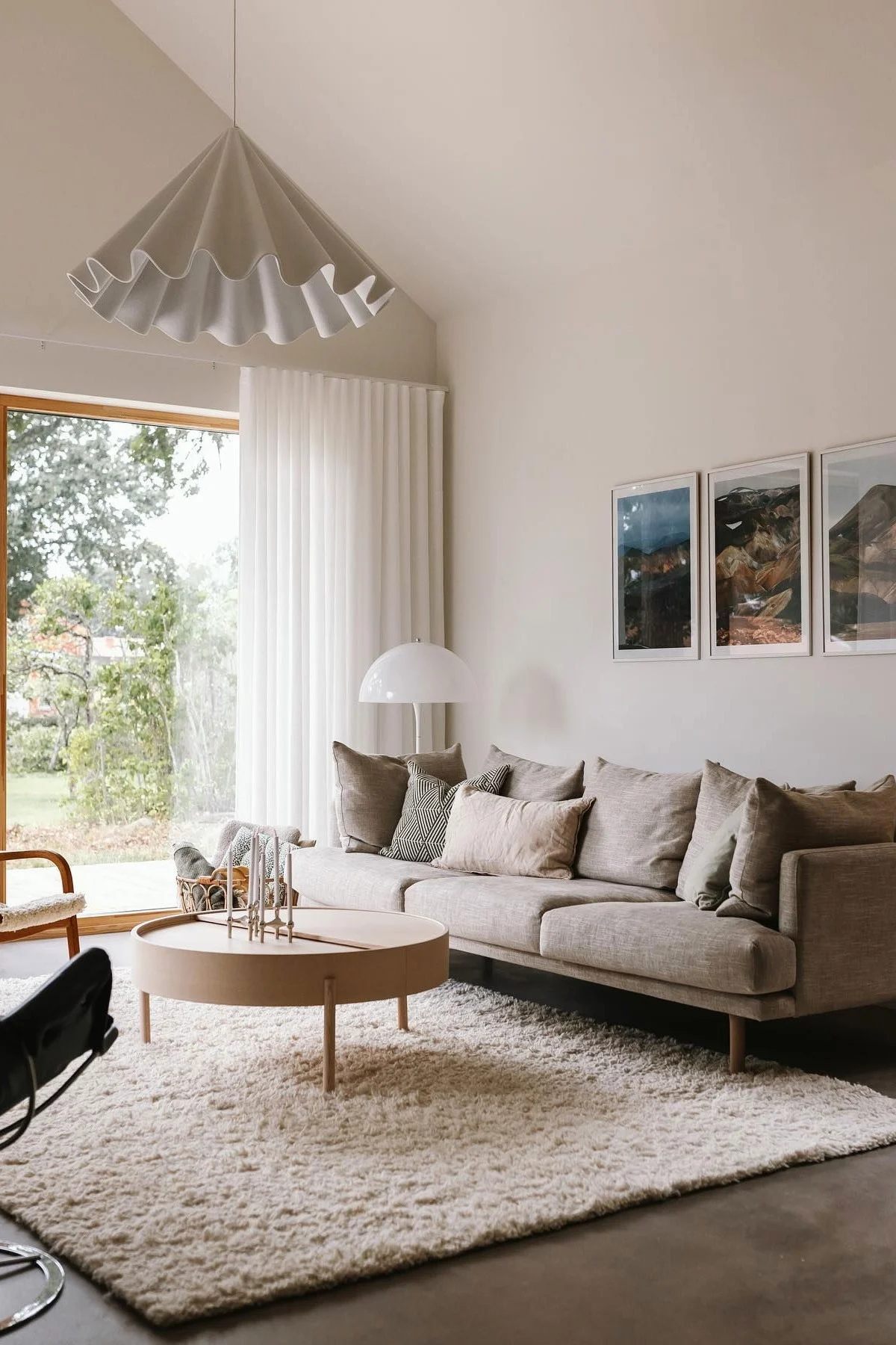Modern living room with a beige sofa, round wooden coffee table, shaggy rug, large window, pleated ceiling lamp, and wall art.