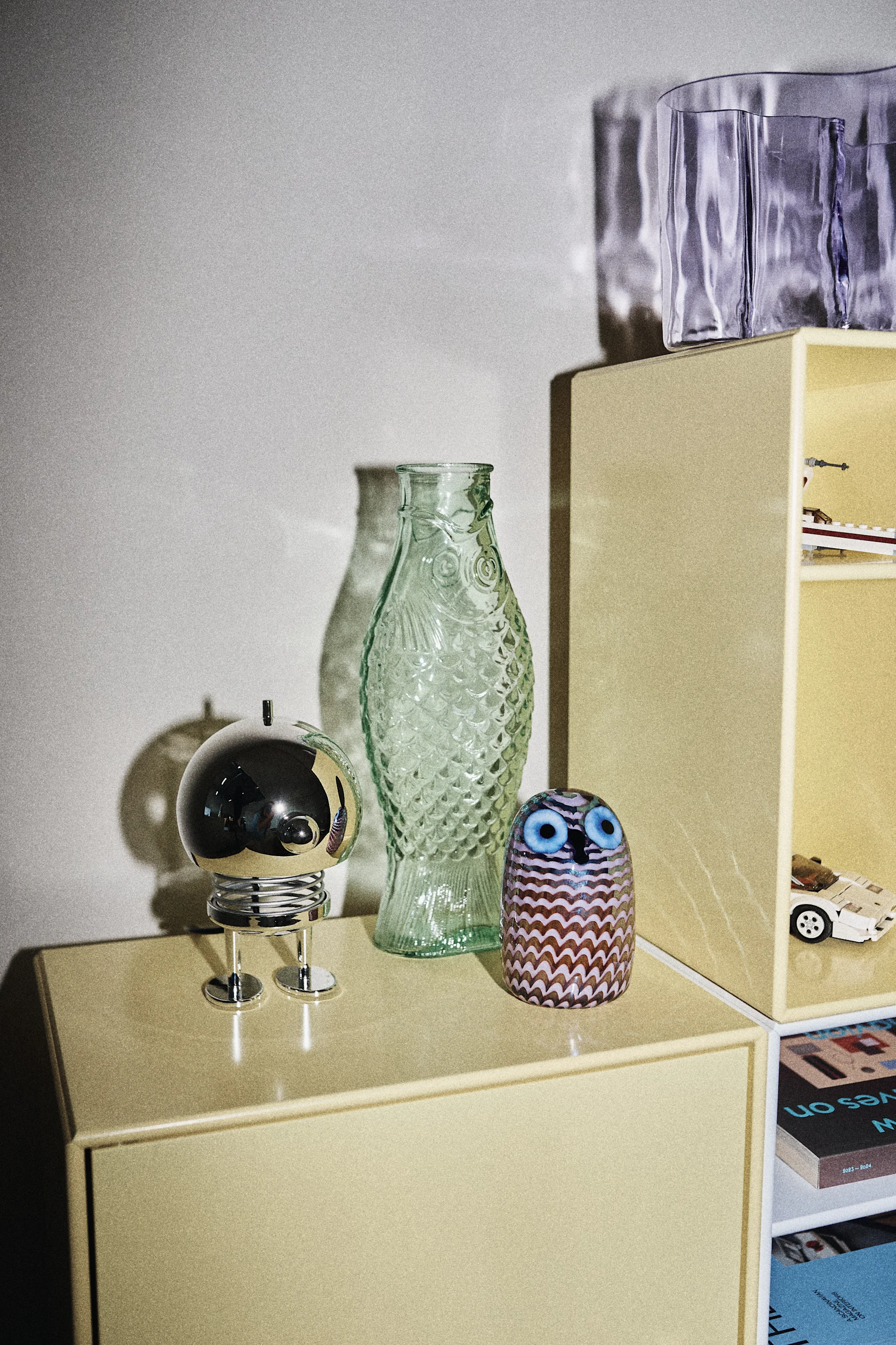 Decorative items on a yellow cabinet: a silver sphere, a green fish-shaped vase, and a patterned glass owl, next to a yellow bookshelf.