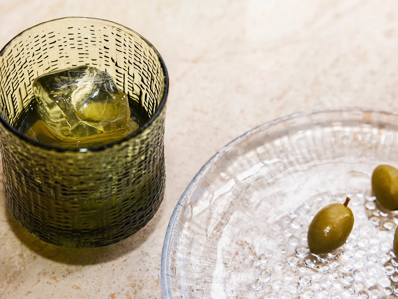 A dark green textured glass with a drink and ice, next to a clear textured plate with green olives.