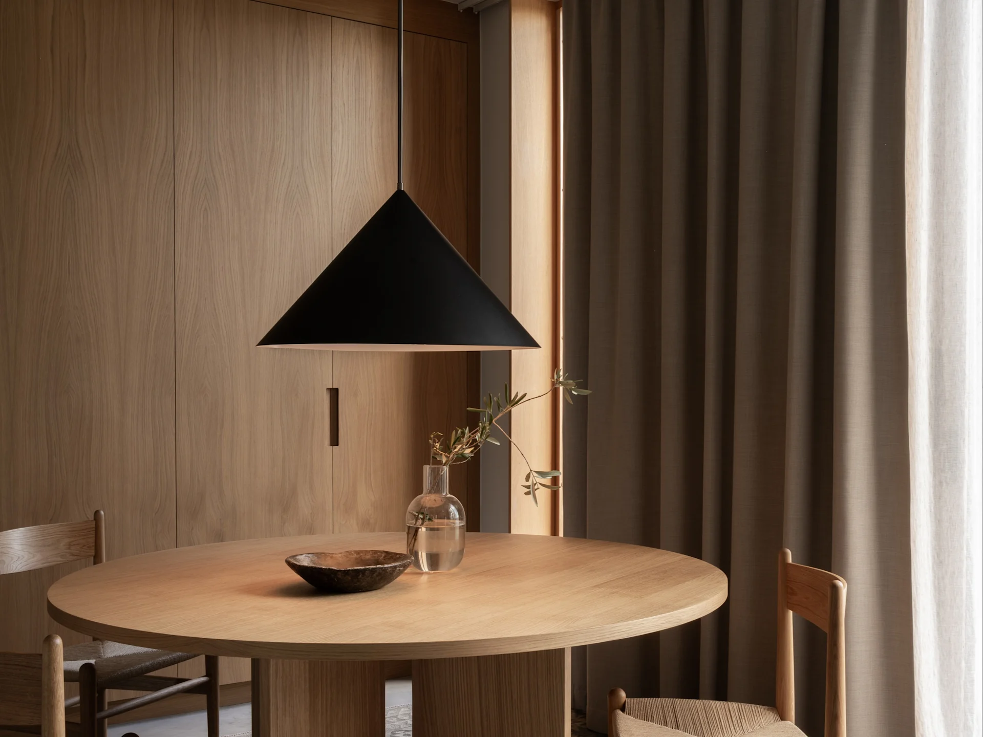 A modern dining space with a round wooden table, black pendant lamp, wooden chairs, and a vase with a branch.