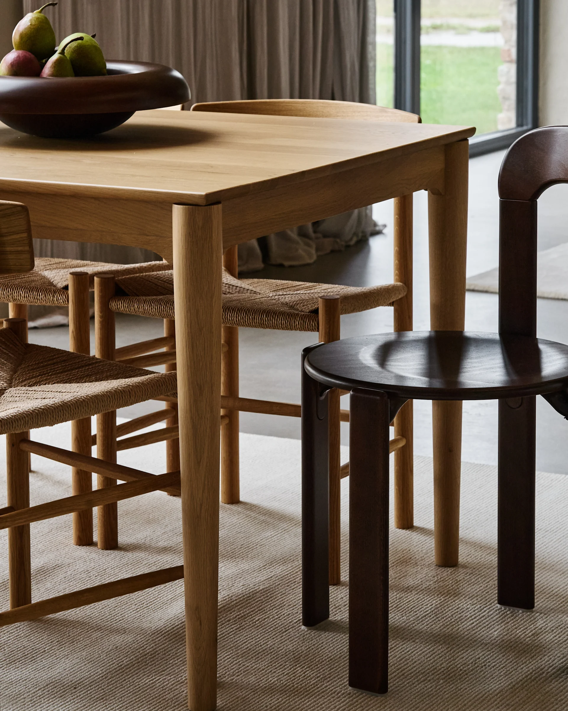 A light wooden dining table with a dark fruit bowl and chairs, two with woven seats and one dark wooden, on a light rug.