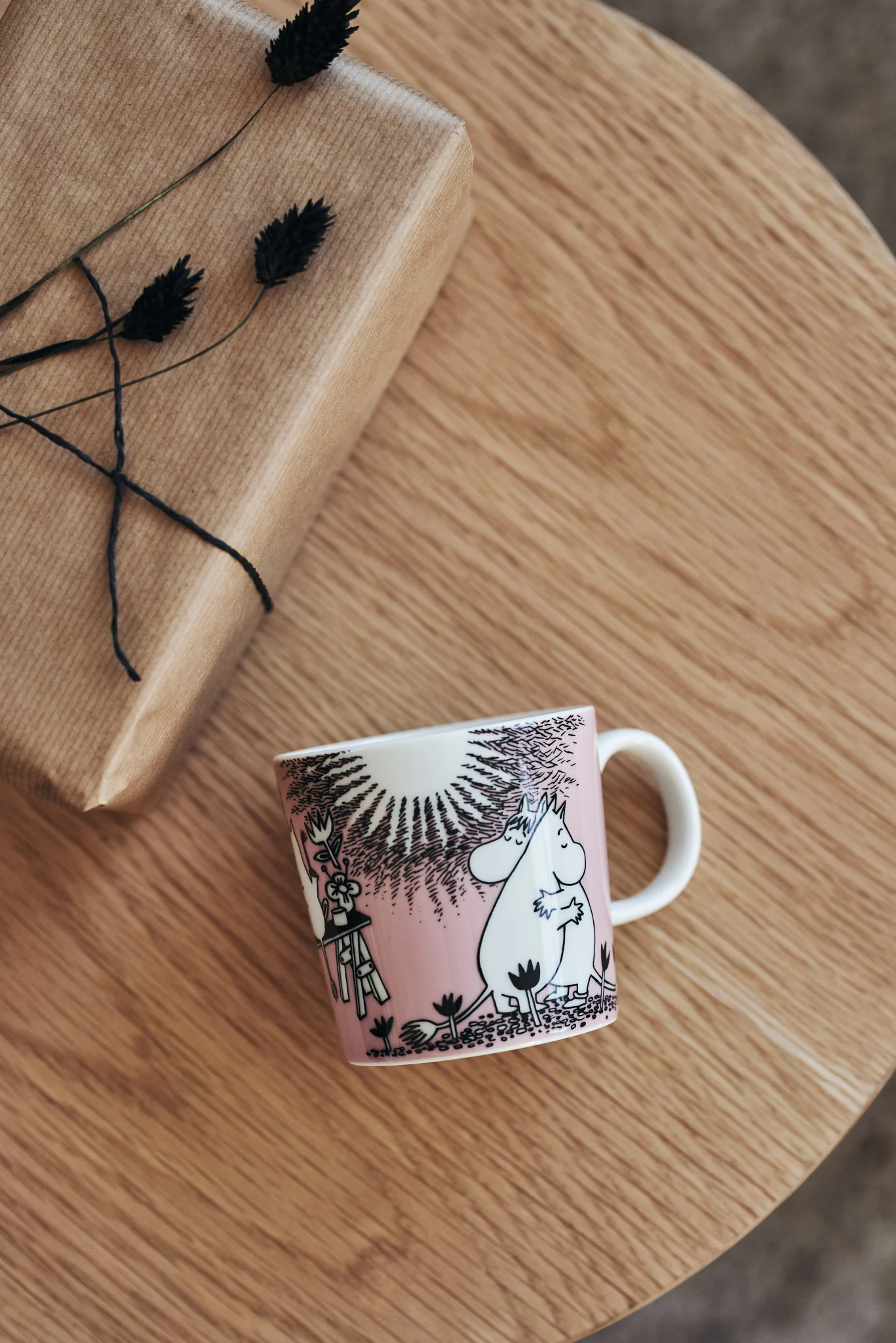 A pink mug with a white hippo-like character illustration and a wrapped gift on a wooden table.