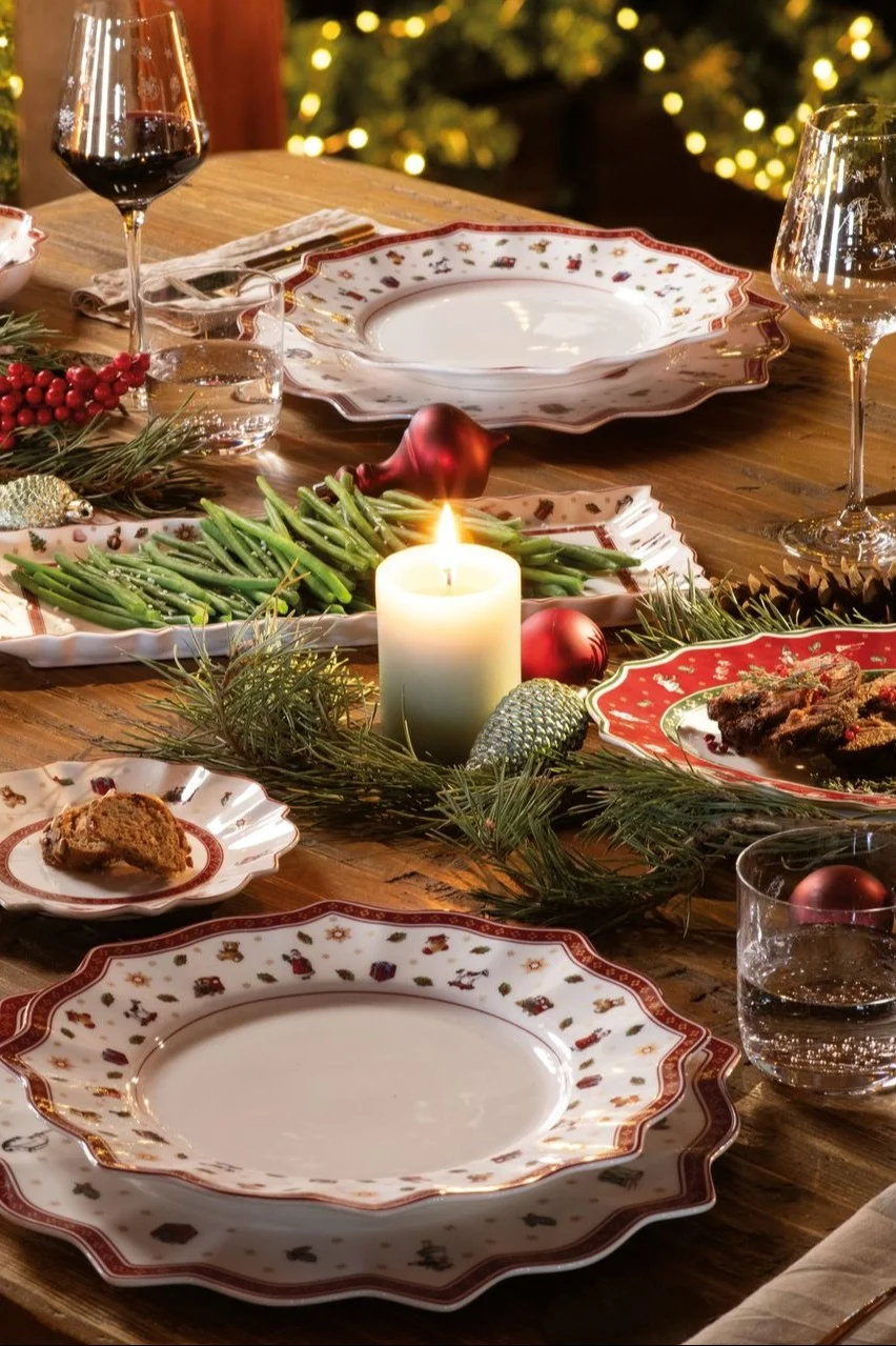 A festive Christmas dinner table set with white patterned plates with a red rim, wine glasses, green beans, a lit candle, and pine garland.