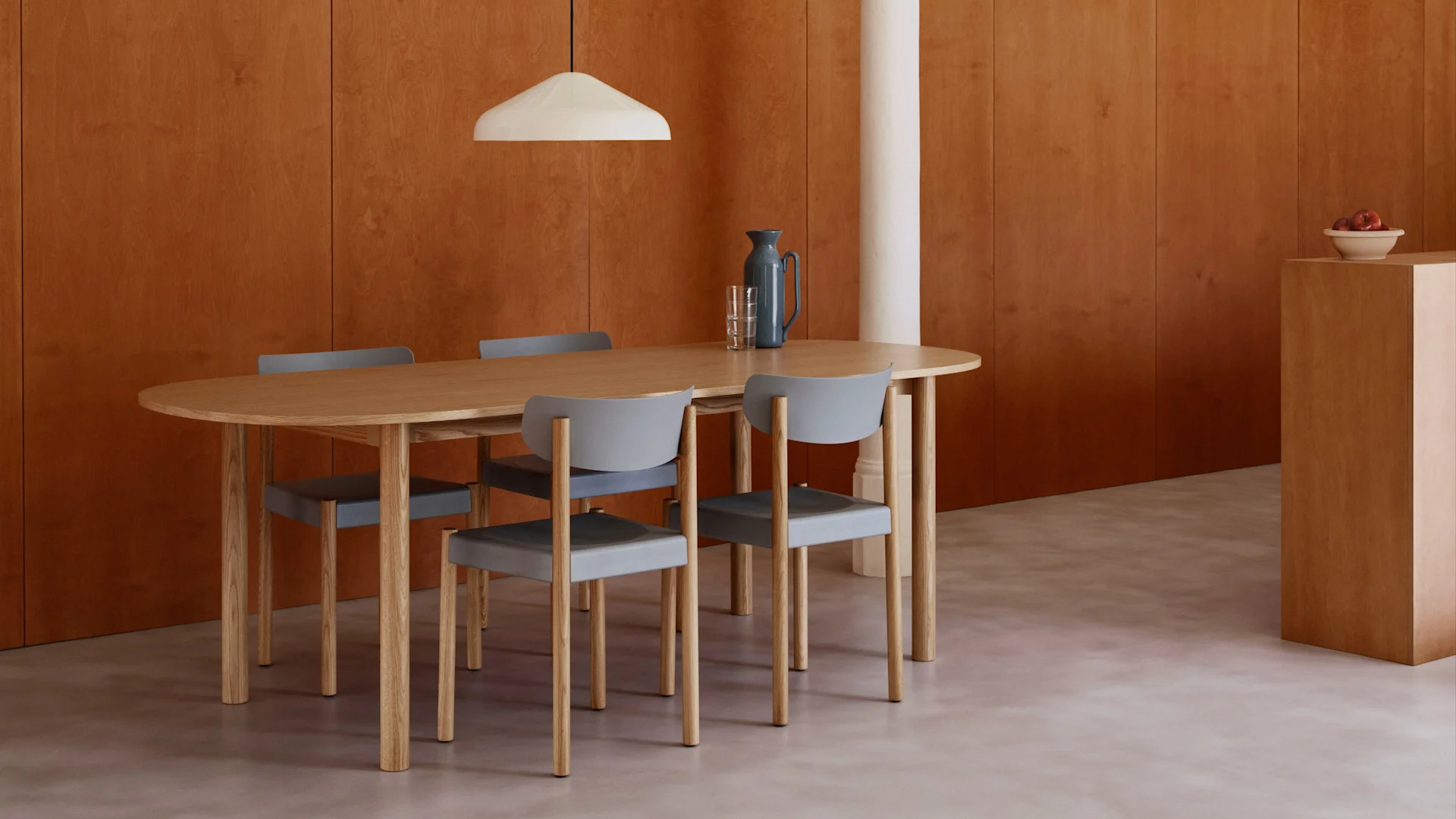 Modern dining room with an oval wooden table, four grey chairs, warm wood-paneled walls, light floor, and a white pendant lamp.