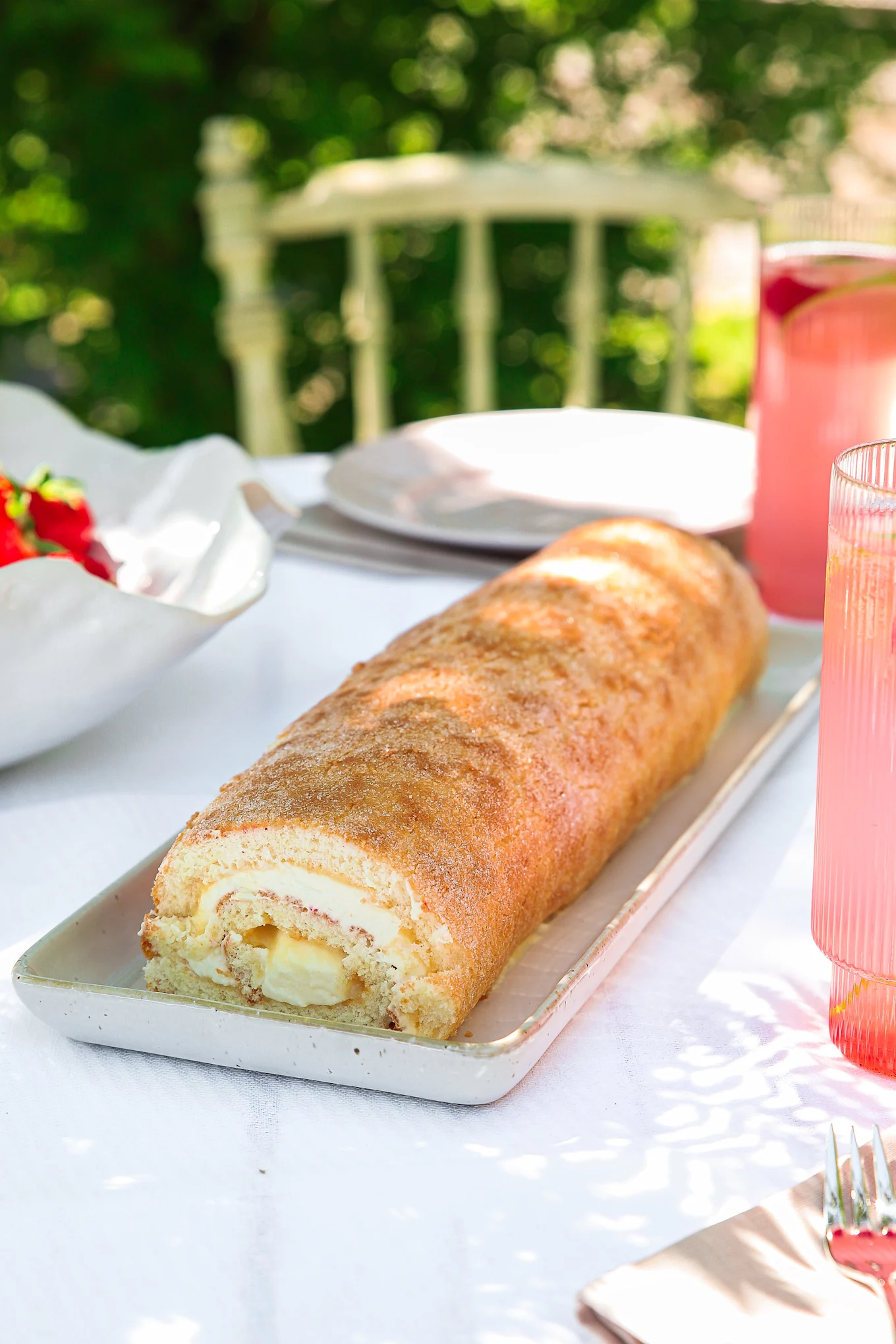 Dessert roll with cream and banana filling, dusted with sugar, served on an outdoor table with strawberries and pink drinks.