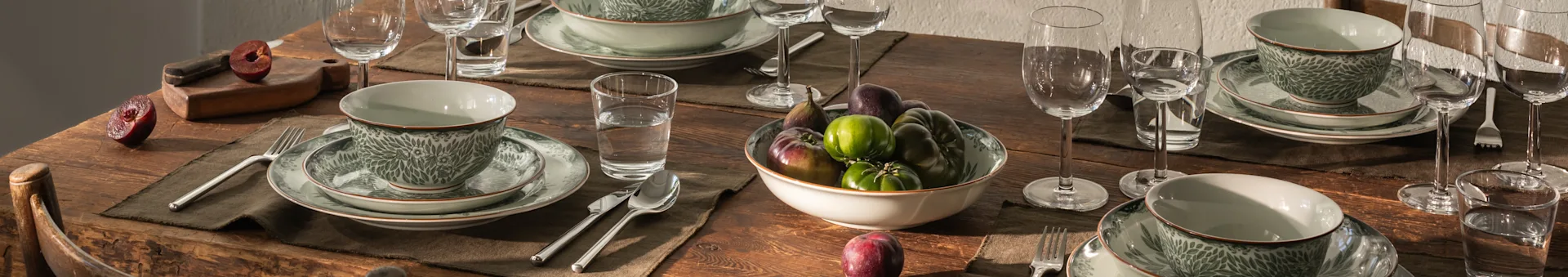 A rustic wooden dining table set with elegant green patterned dinnerware, clear glasses, and silverware, with plums.