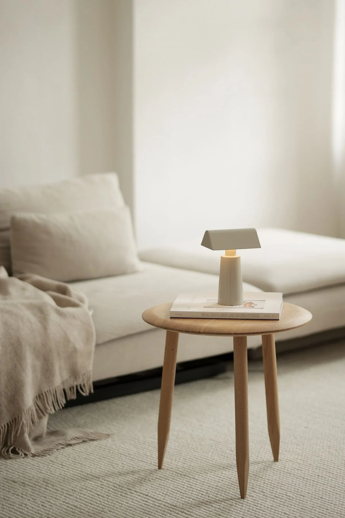 A minimalist light gray portable lamp with a rectangular shade on a round light wood side table next to a beige sofa with a throw.
