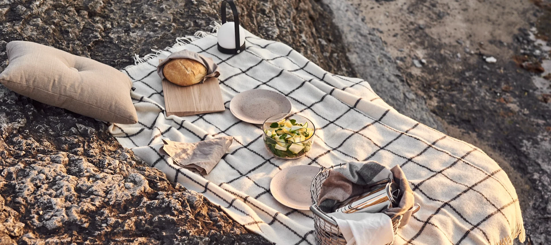 Picnic on rocks: plaid blanket, beige pillow, bread, lantern, salad, plates, and a basket with a blanket and cutlery.