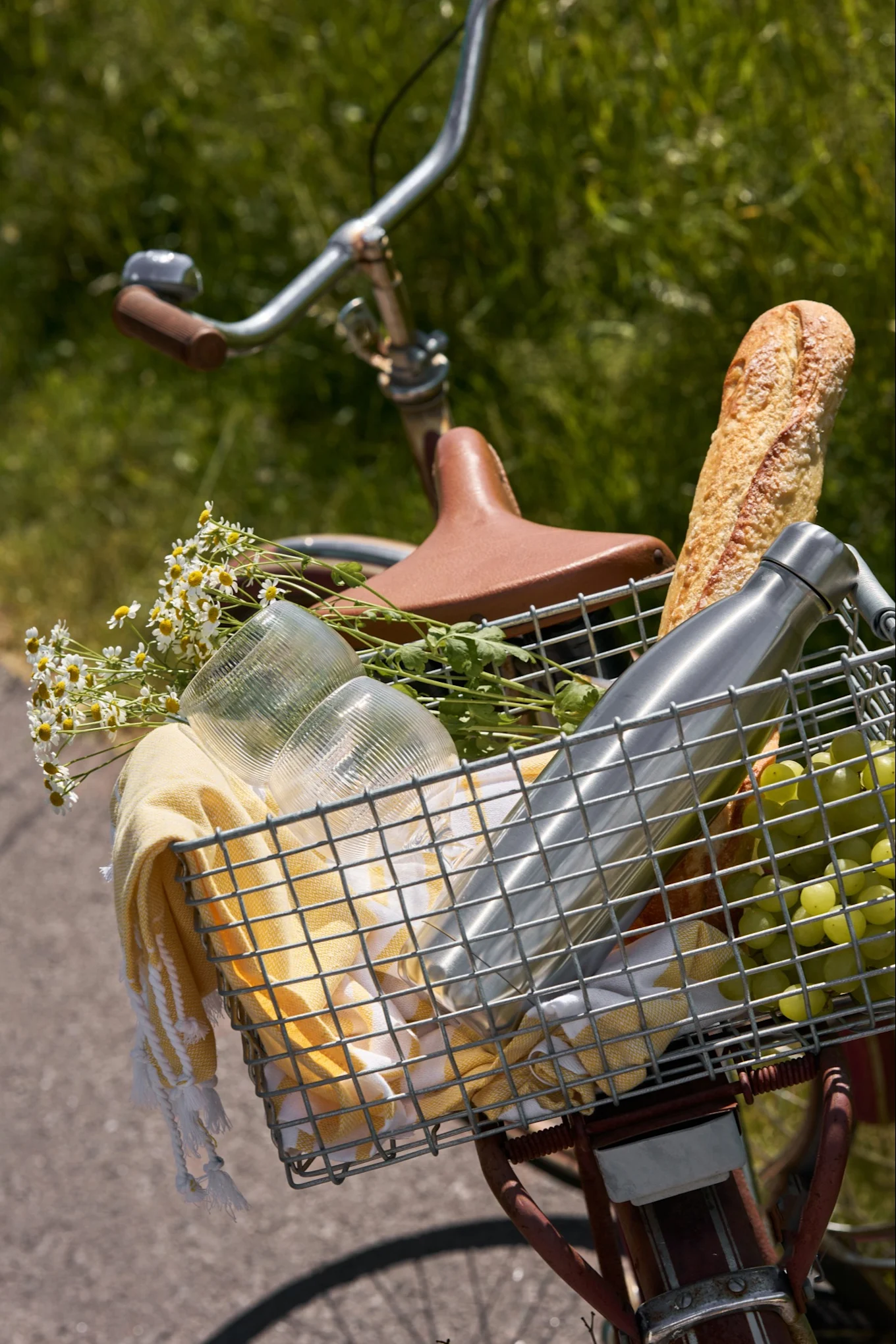 A bicycle's front basket filled with picnic items: a baguette, water bottle, grapes, cups, a yellow towel, and white daisies.