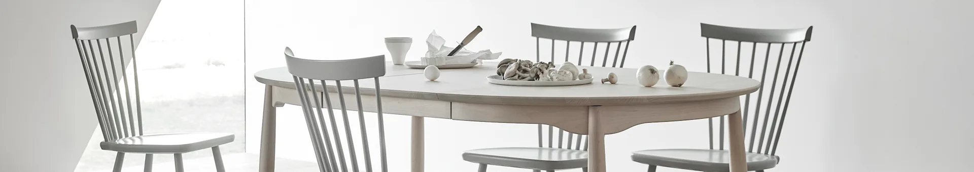 Oval light wood dining table with four grey spindle chairs, set with a plate of mushrooms, an egg, cups, and cheese.