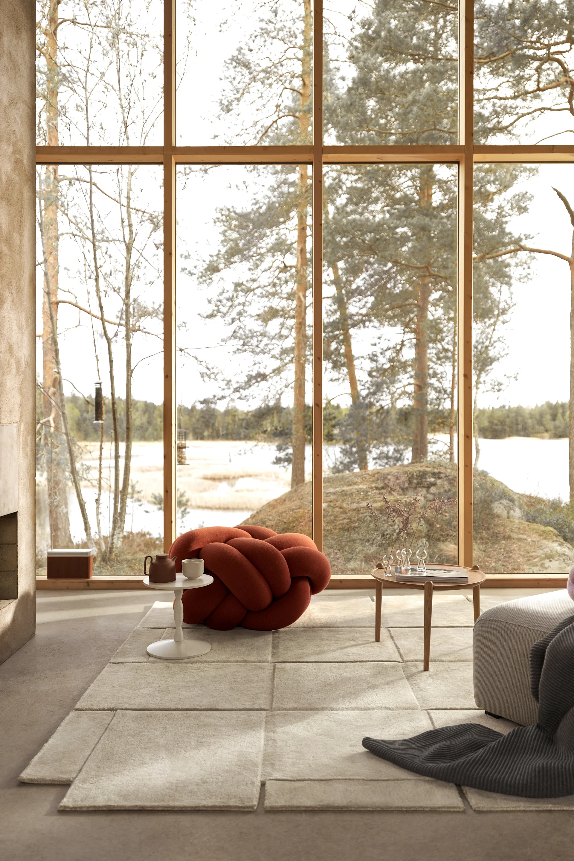 Modern living room with large windows overlooking a lake. Features a rust-orange knot ottoman, a beige rug, and wood furniture.