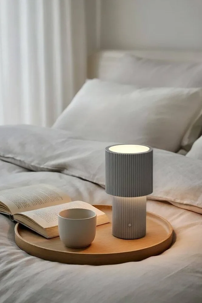 A gray, fluted portable lamp, an open book, and a white mug on a wooden tray on a light-colored bed.