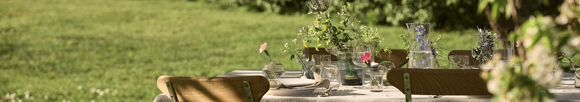 Outdoor dining table set with white linen, wooden chairs, and various fresh flower arrangements in glass vases on a sunny lawn.