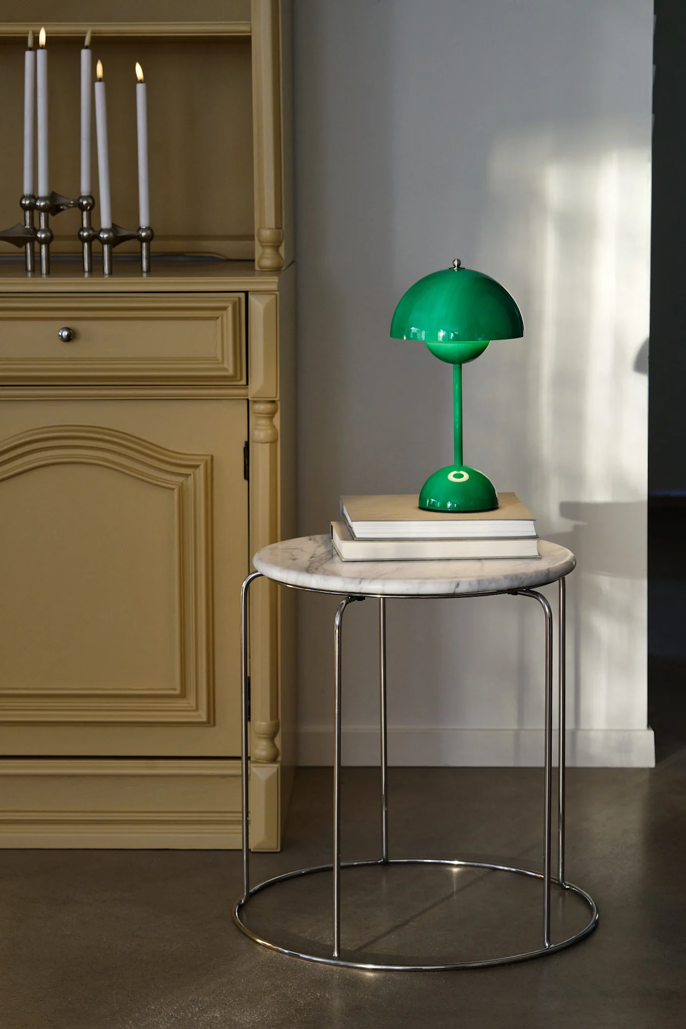 A green mushroom-shaped table lamp sits on books on a round marble table, next to a yellow cabinet with lit candles.