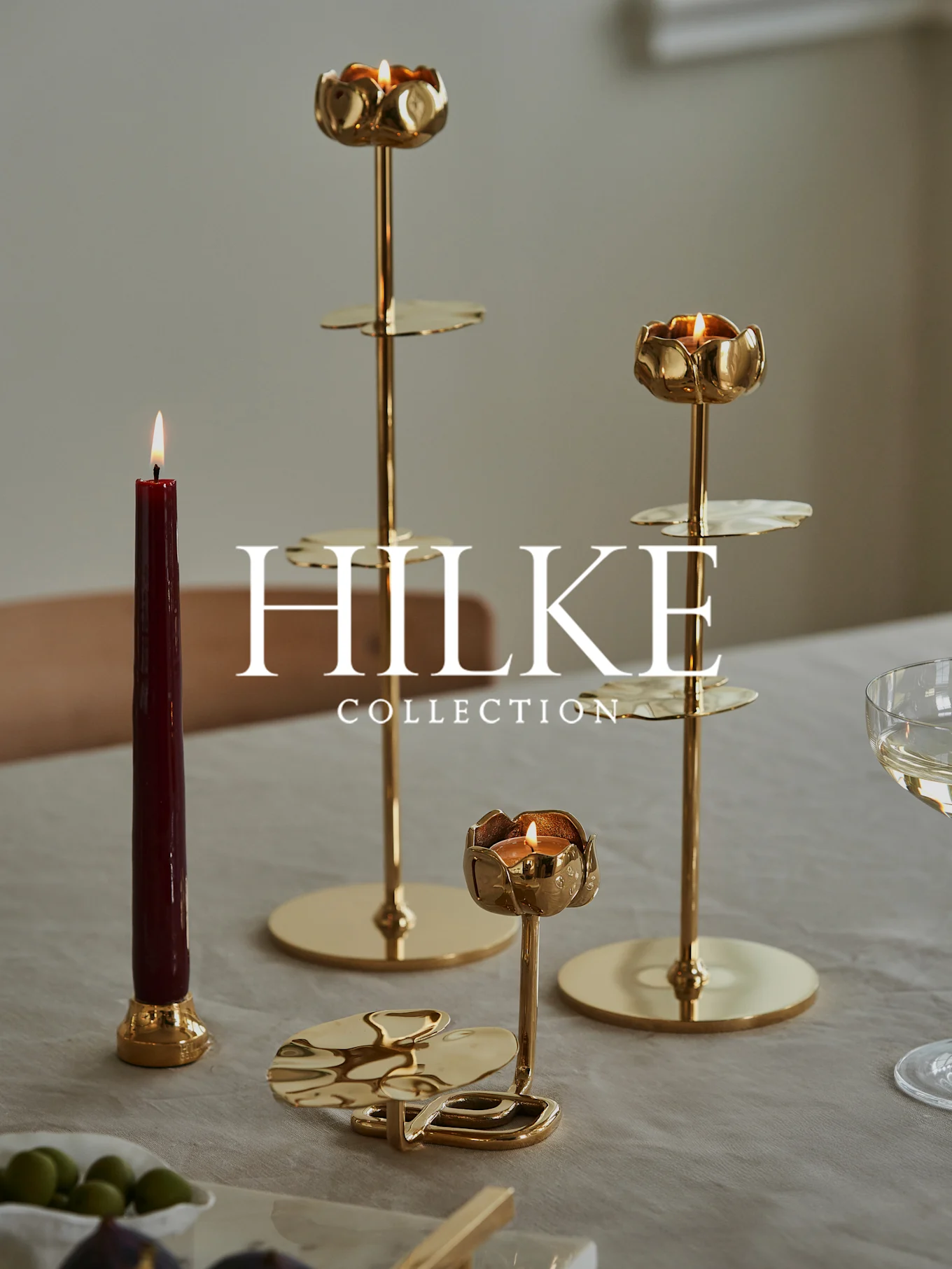 Three gold floral candle holders with lit candles and a red taper candle on a table.
