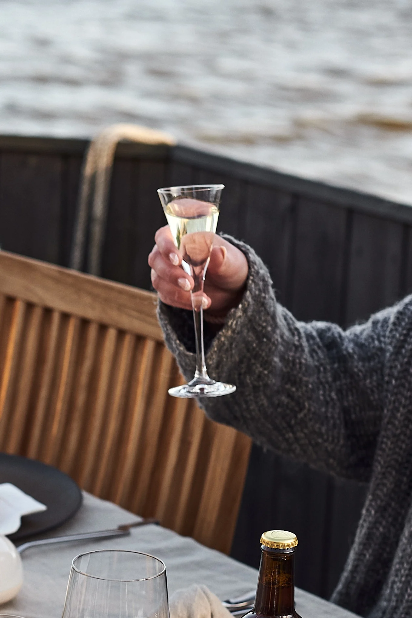 A hand in a grey sweater holds up a delicate glass of white wine or champagne at a table set outdoors by water.