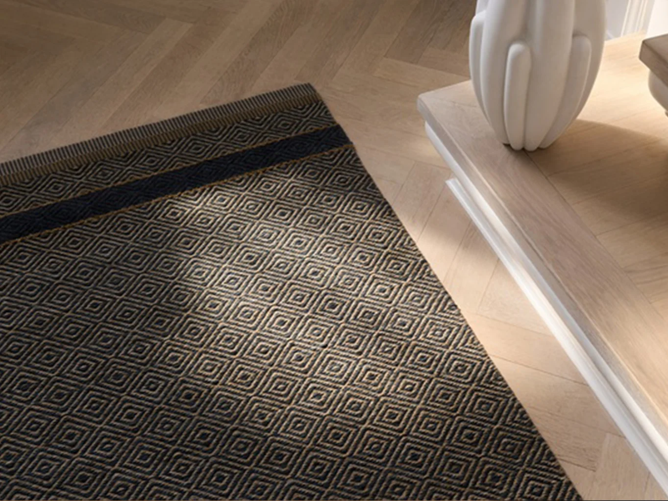Dark blue and natural fiber rug with diamond pattern on a herringbone wooden floor, next to a table with a white vase.