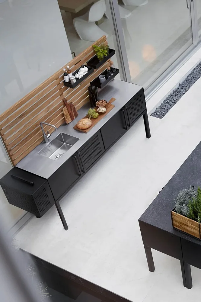 Modern outdoor kitchen with a black unit, stainless steel sink, wooden back panel, food on a cutting board, and a herb planter.