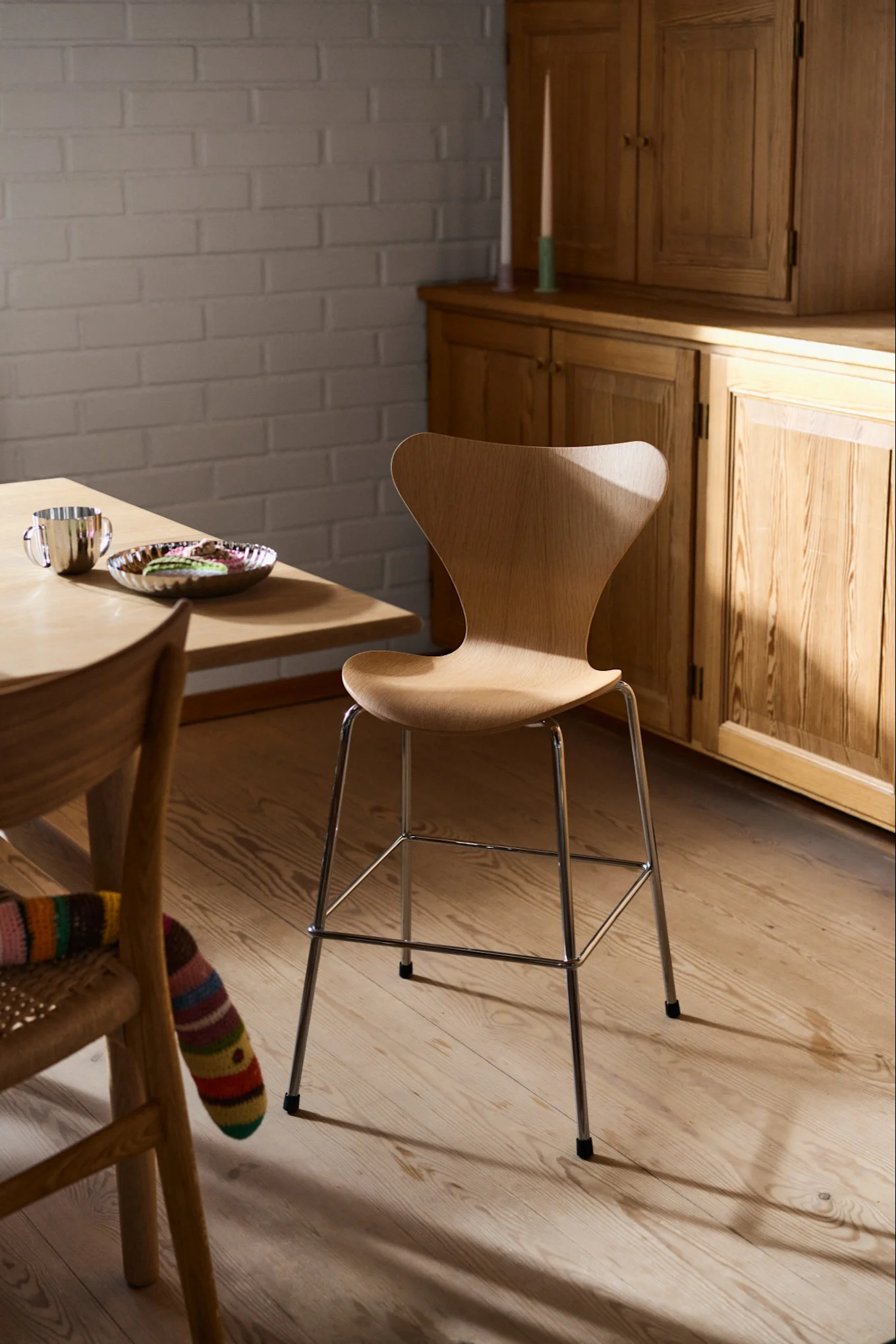 Modern wooden stool with chrome legs in a sunlit room, next to a table with a cup and pastries.