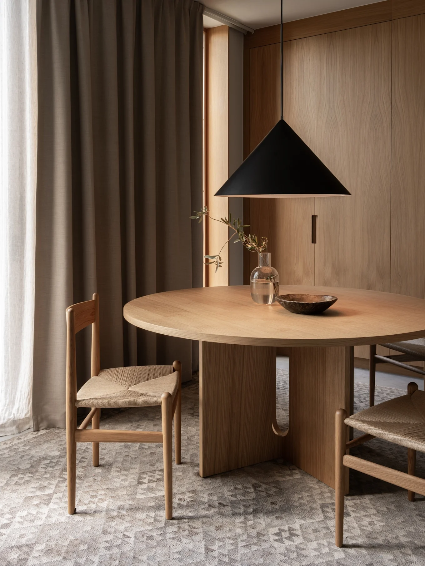 A minimalist dining area with a round wooden table, two chairs with woven seats, a black pendant light, and wooden walls.