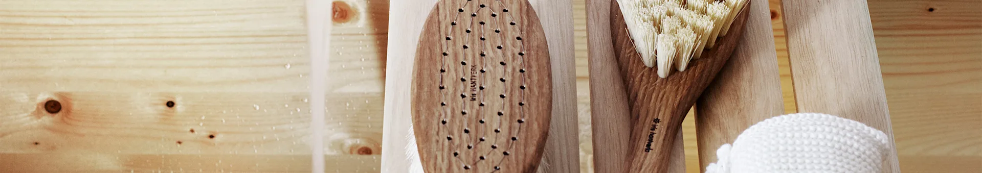 A wooden dry brush, a bath brush with natural bristles, and a white woven scrubber on a wet wooden surface.