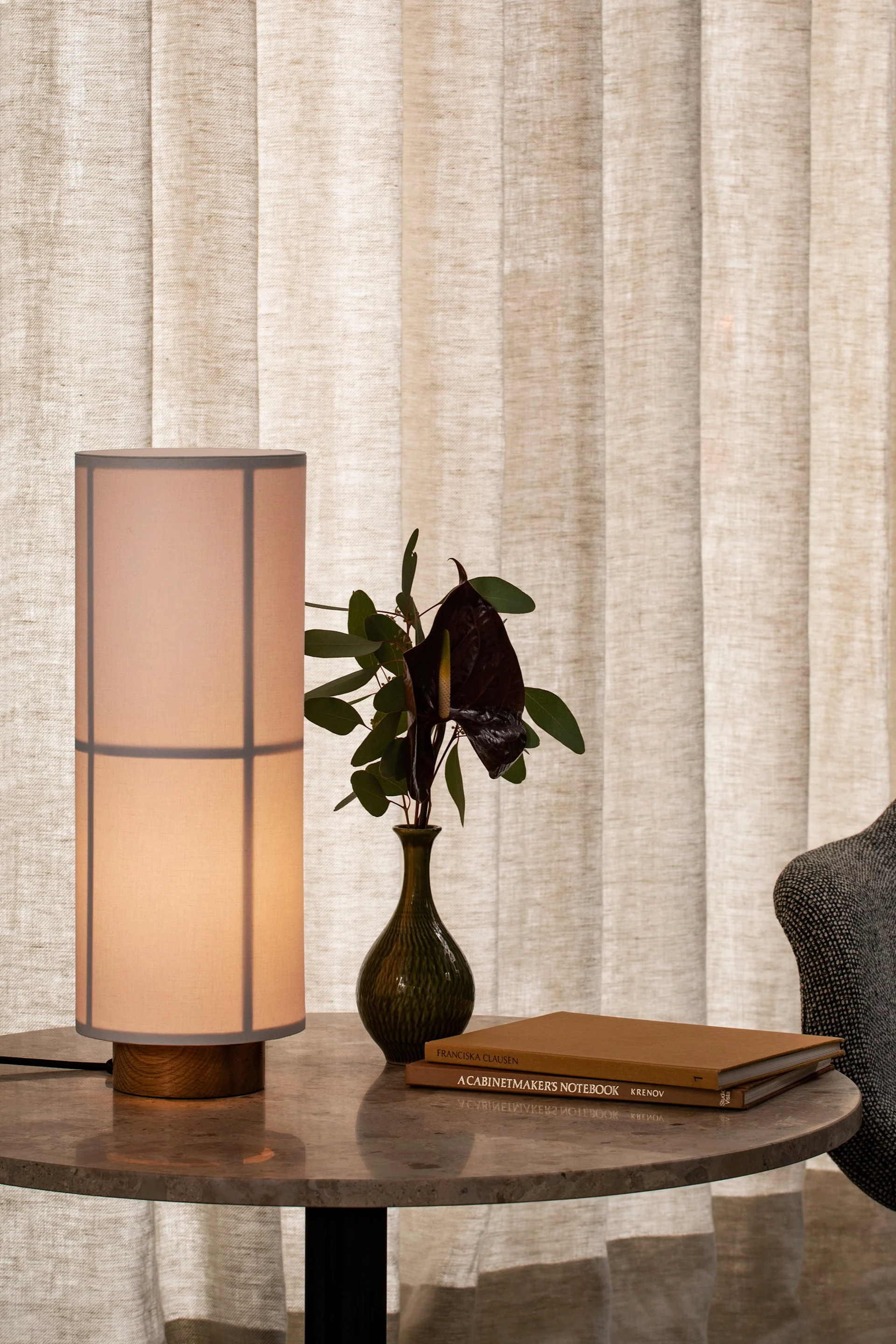 A lit table lamp, dark vase with flowers, and books on a marble table. Light fabric curtains in background.