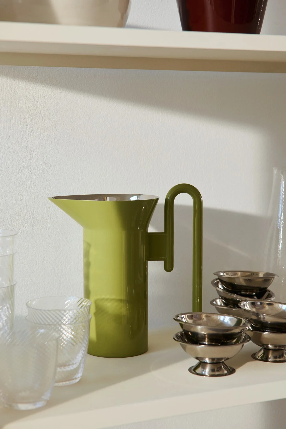 Olive green pitcher with a U-shaped handle, flanked by clear patterned glasses and stacks of small silver dessert bowls on a shelf.