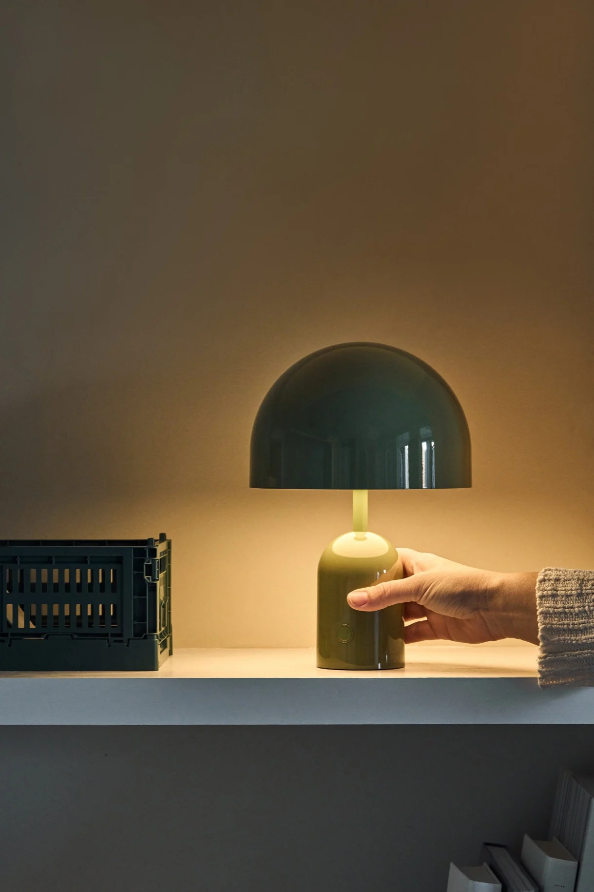 A hand adjusts a glowing dark green mushroom-shaped table lamp on a white shelf next to a matching storage crate.