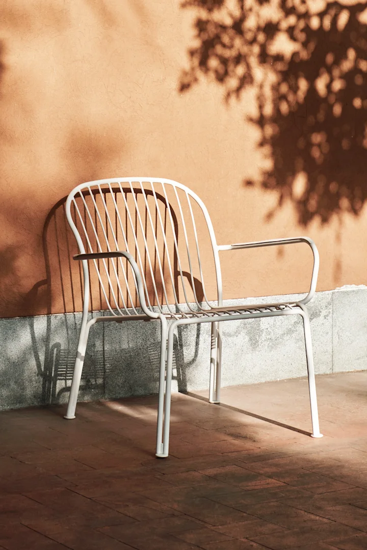 White slatted outdoor chair against an orange wall, with shadows of leaves and the chair on the wall and floor.