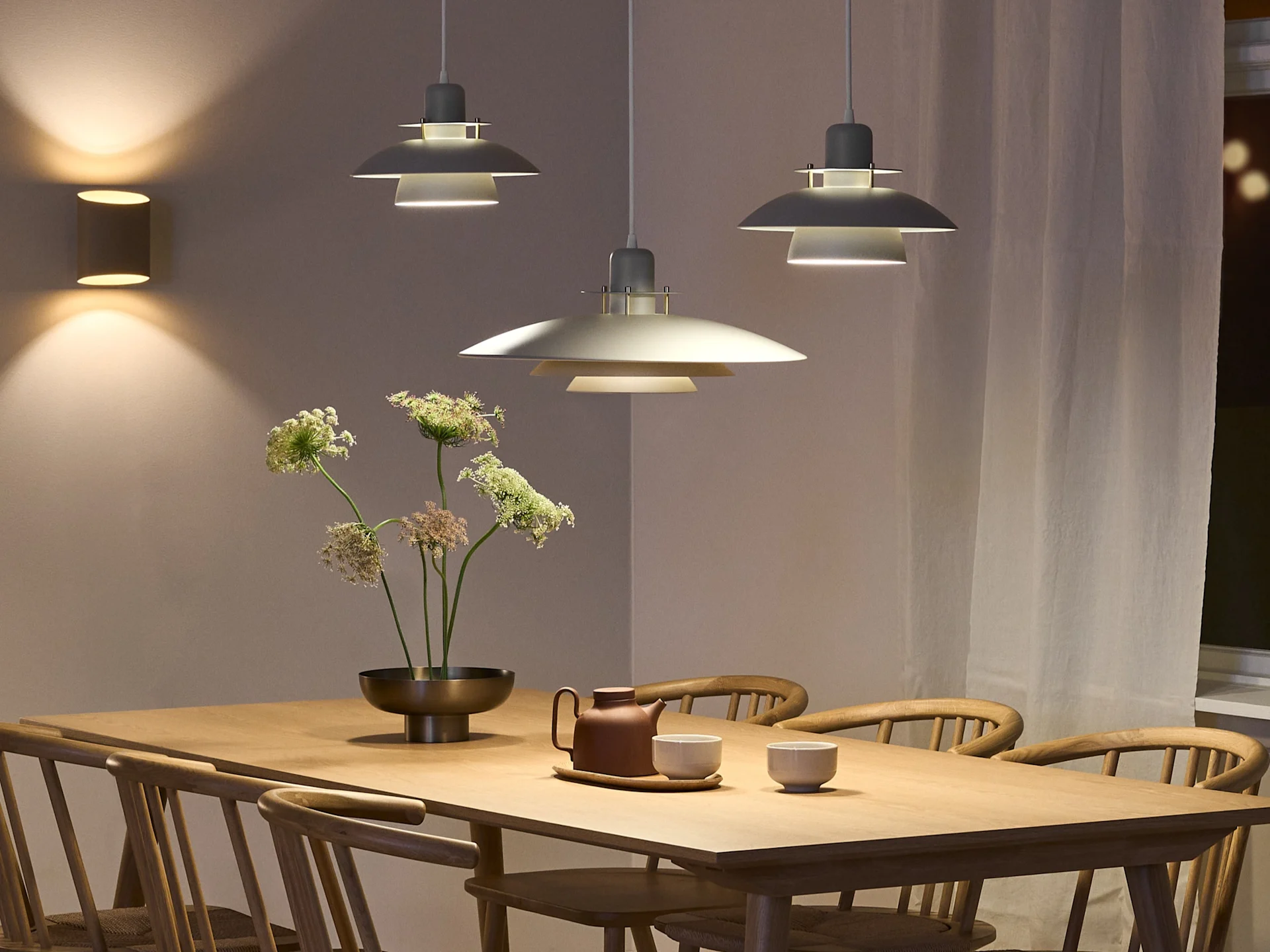 A dining room with a wooden table, chairs, and three grey pendant lights. A wall sconce lights the wall, with flowers, a teapot, and cups on the table.