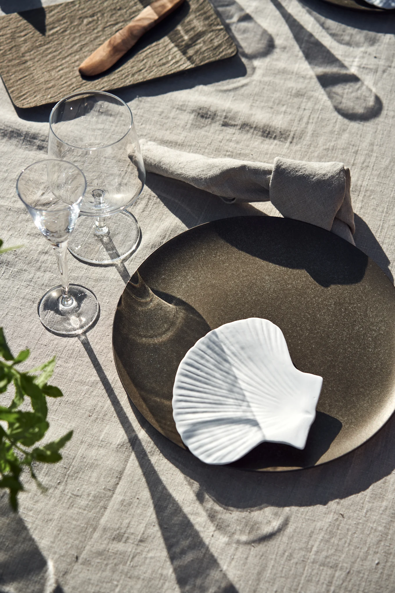 A single place setting with a dark dinner plate, a white seashell dish, two wine glasses, and a linen napkin on a light tablecloth, with strong shadows.