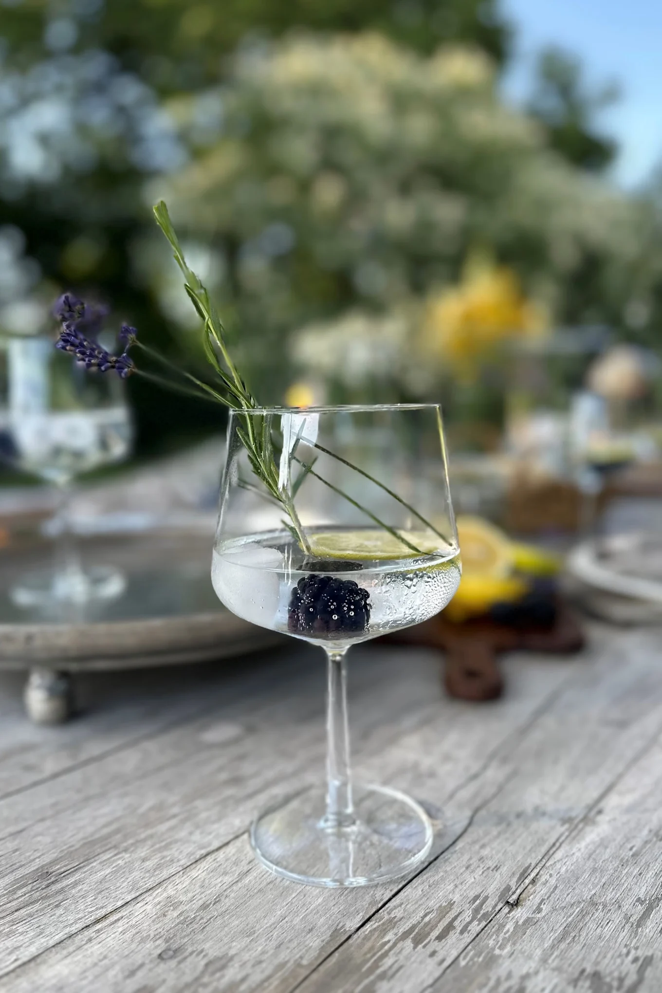 A clear stemmed glass on a wooden table, containing a blackberry, a lime slice, a lavender sprig, and clear liquid.