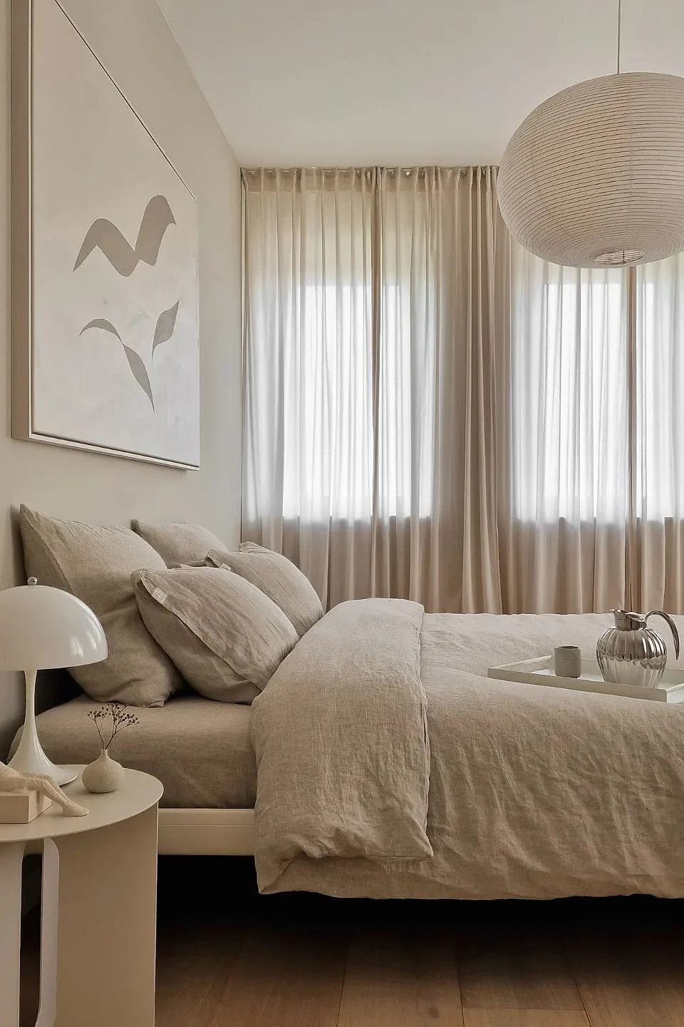 A minimalist bedroom with a bed featuring light beige linen bedding, a mushroom lamp, and sheer curtains. Abstract art hangs above the bed.