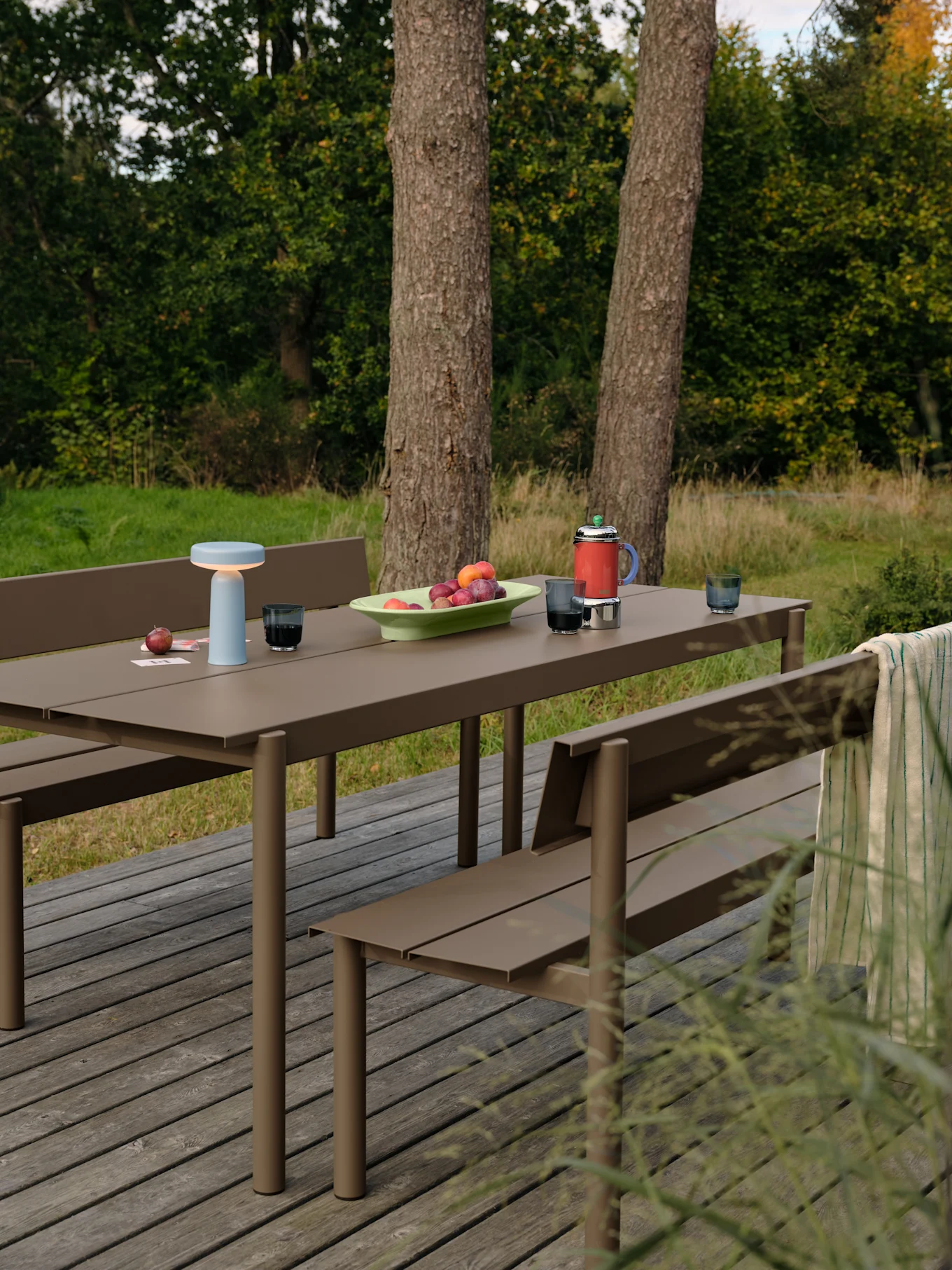Outdoor brown picnic table and benches on a wooden deck with a light blue lamp, fruit, coffee press, and glasses.