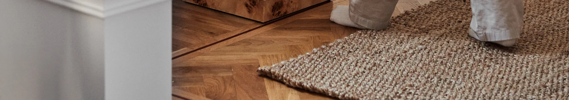 Person's feet on a textured light brown rug over a wooden herringbone floor.
