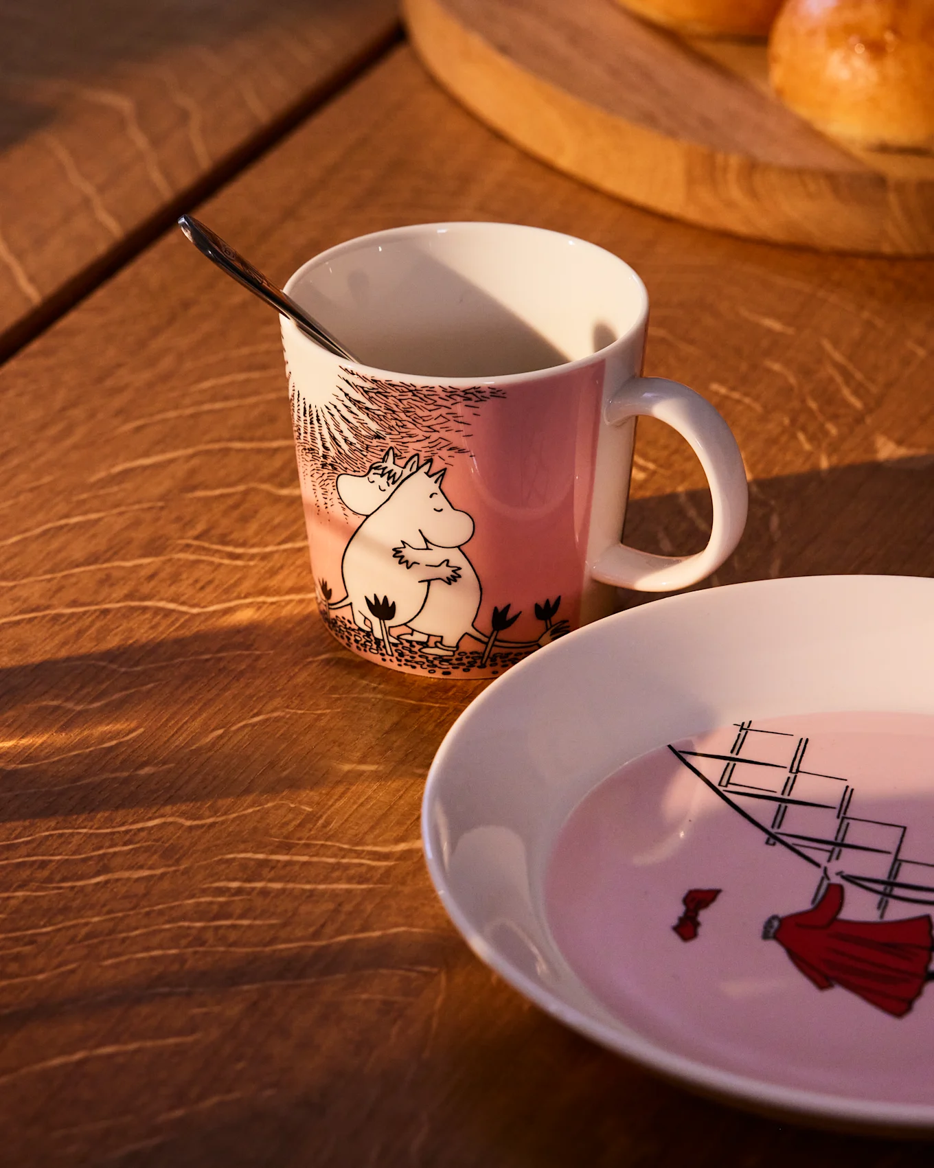 Pink Moomin mug with Moomintroll and Snorkmaiden, next to a matching plate with a sailing ship, on a wooden table.