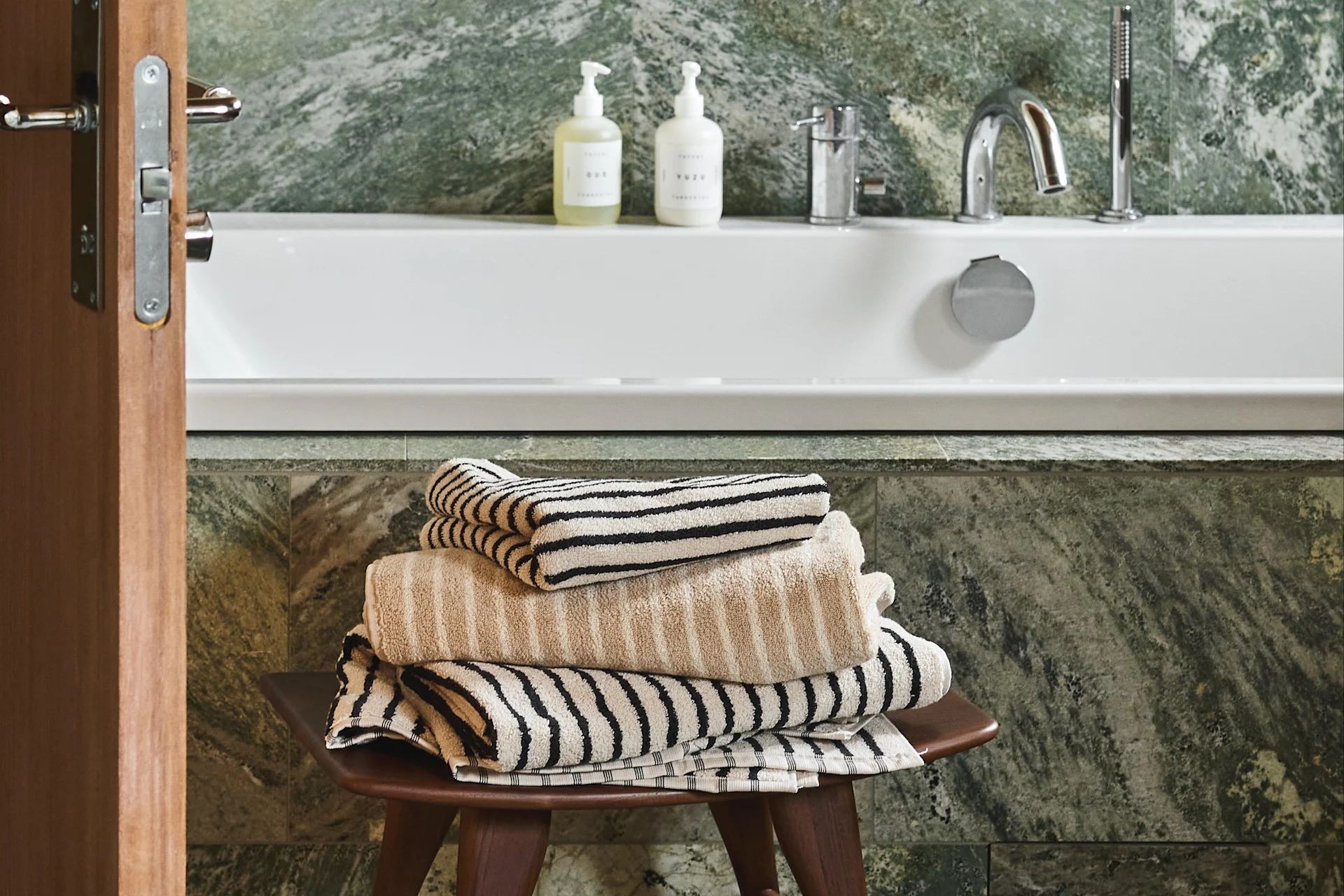 A stack of beige and striped towels on a wooden stool in a bathroom with a white tub and green marble walls.
