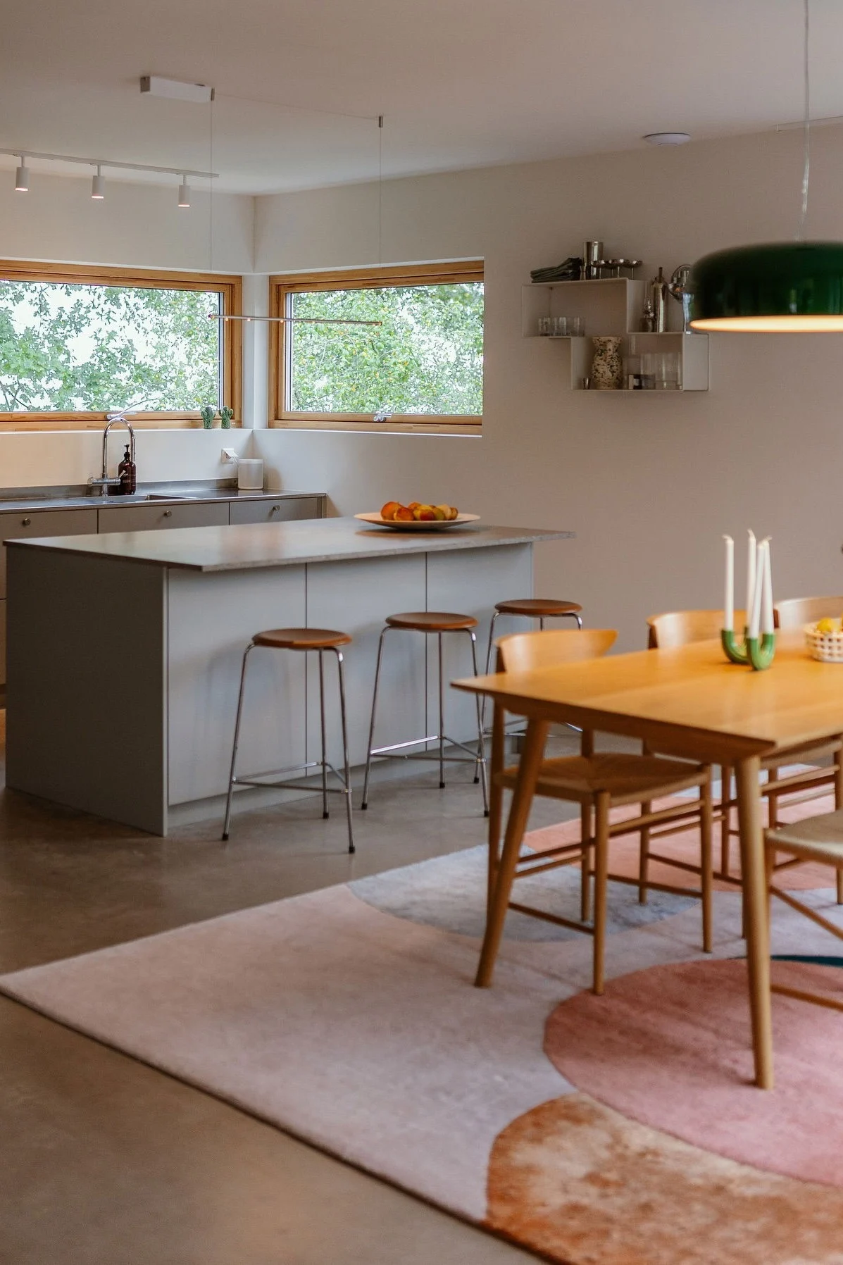 Open-plan kitchen and dining area with a grey island, three stools, a wooden dining table, and a large green pendant lamp.