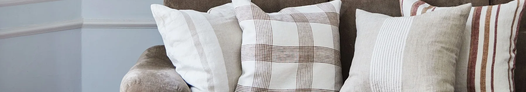 Close-up of three pillows on a brown sofa, featuring white, beige, and plaid patterns with brown and red stripes.