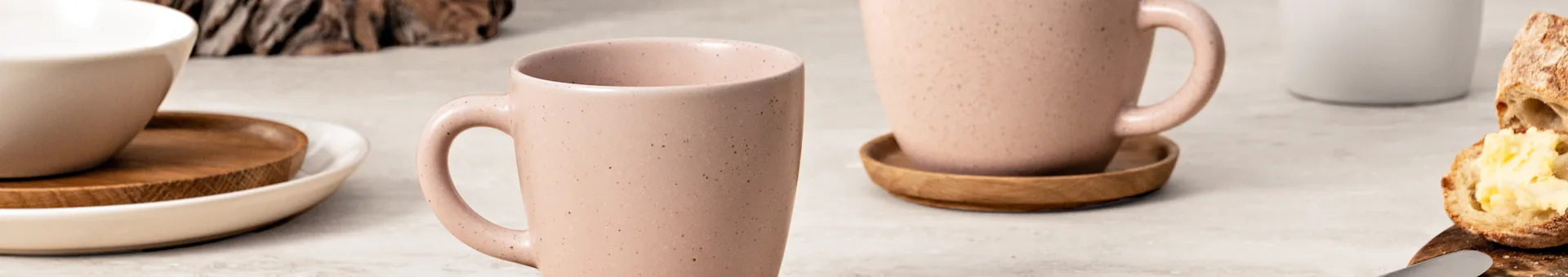 Two speckled pink mugs, a white bowl, and a buttered bread slice on a light background.
