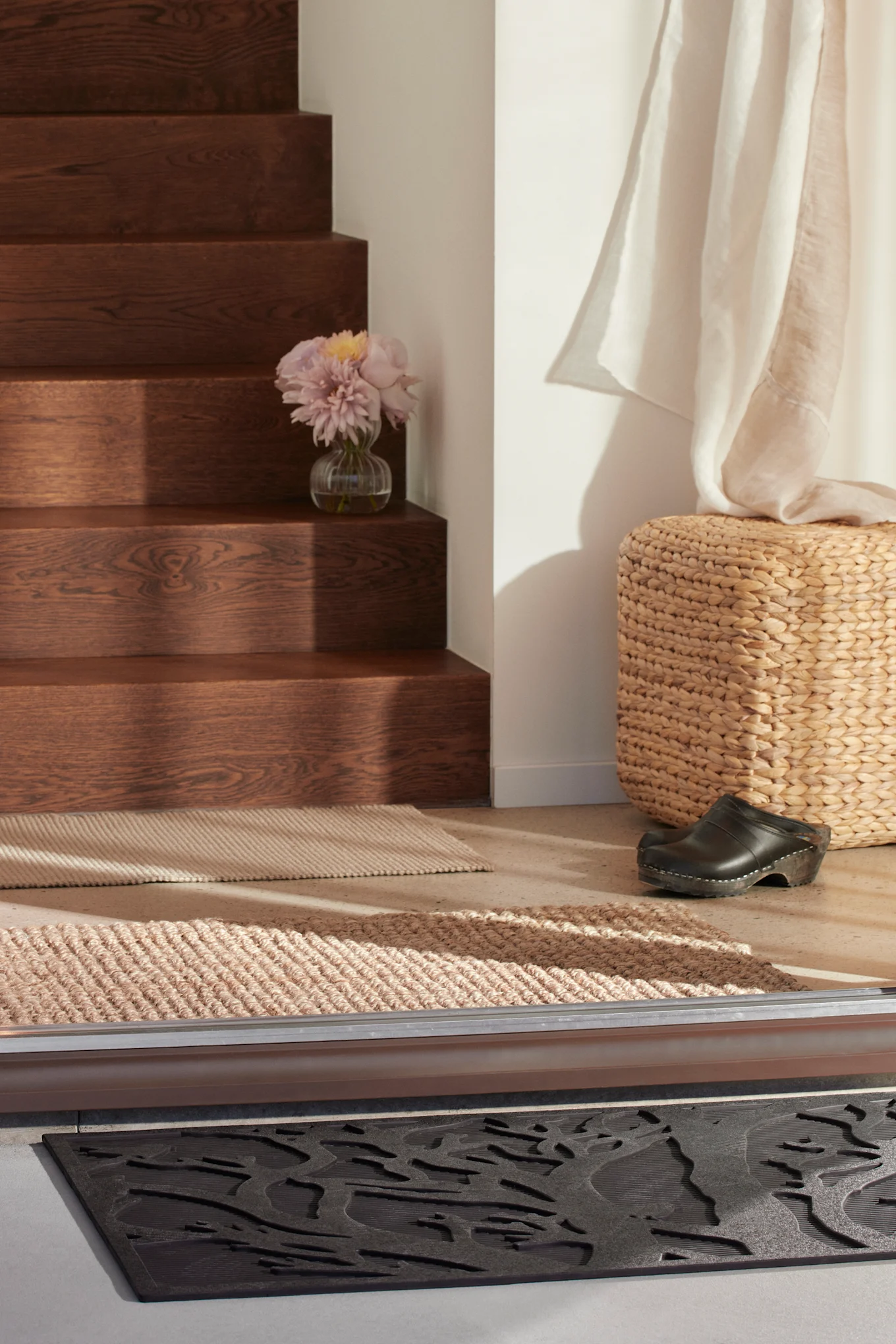 An entryway with dark stairs, a vase of flowers, two beige rugs, a woven pouf, a black clog, and a dark grey textured doormat.