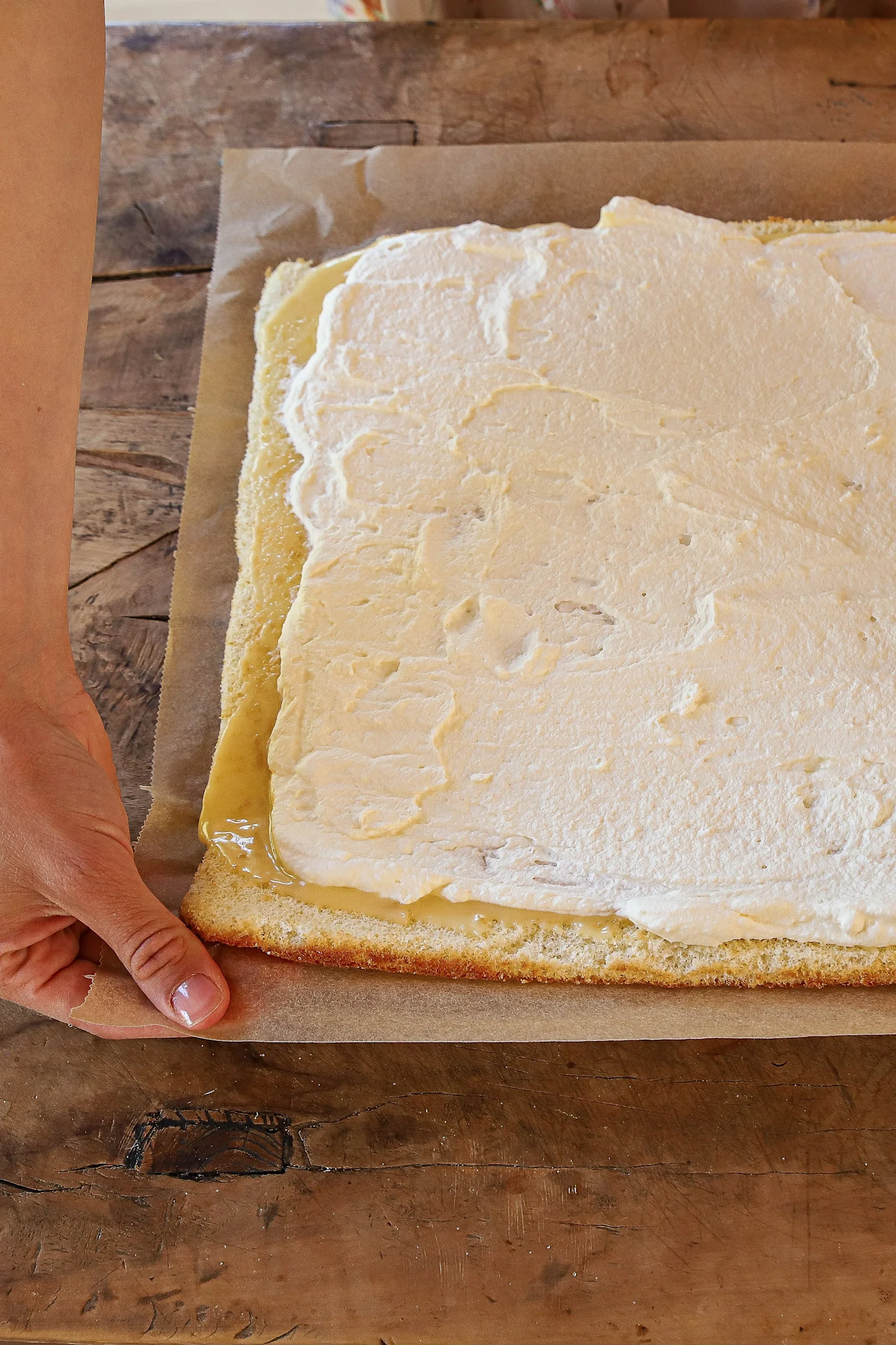 A hand holds parchment paper under a sponge cake base topped with yellow and white cream fillings on a wooden table.