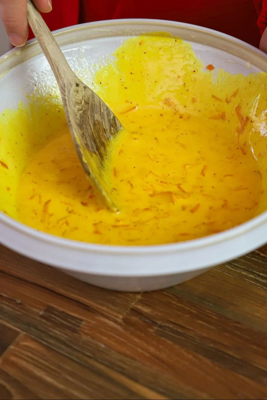 A wooden spoon stirs a yellow batter with orange specks in a white bowl.
