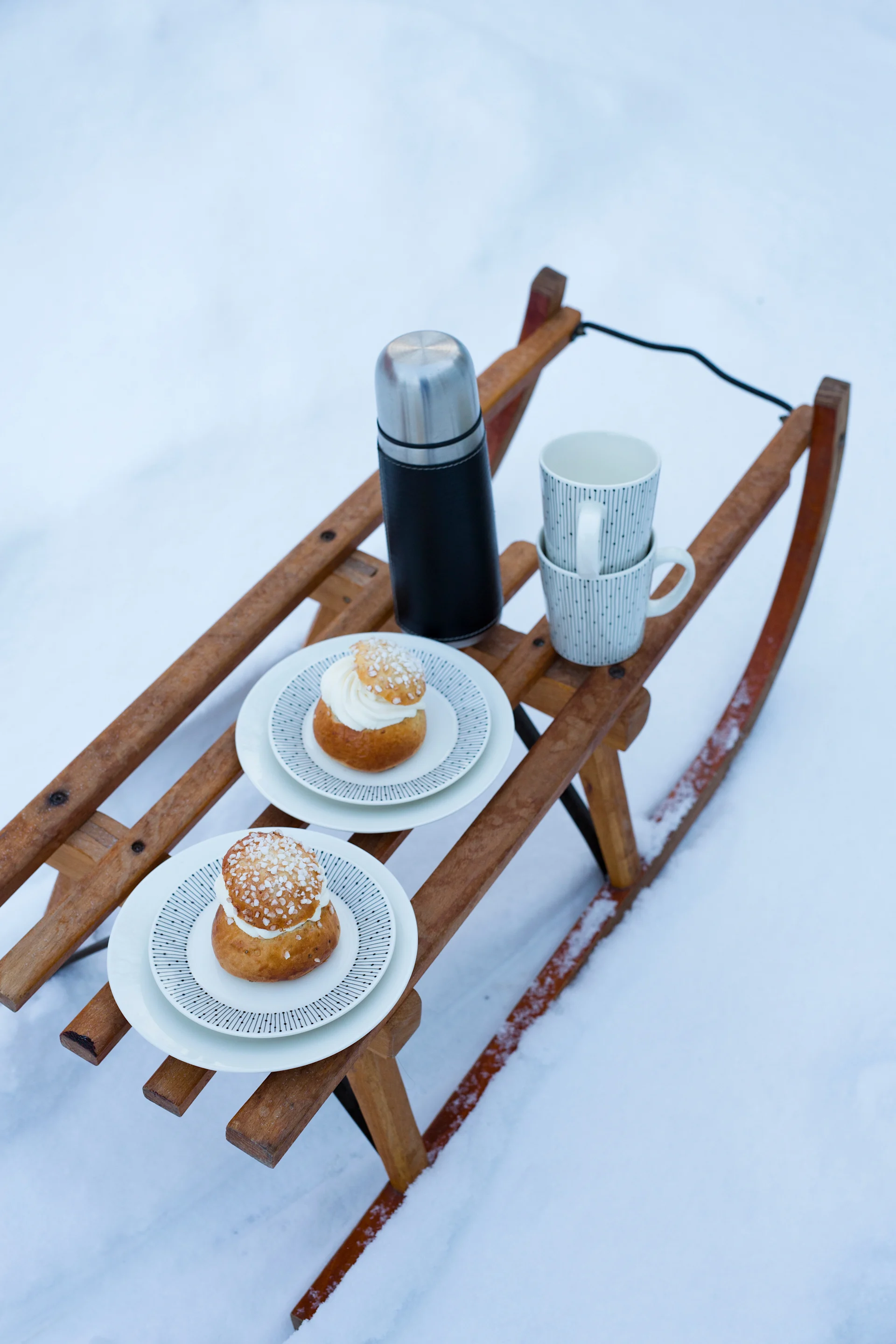 Two cream buns and hot drinks on a wooden sled in the snow.